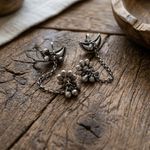 Load image into Gallery viewer, Pair of oxidized silver earcuffs featuring floral motifs, link chains, and small hanging bells on a rustic wooden background.