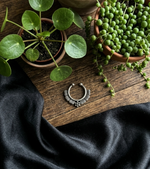 Load image into Gallery viewer, Oxidized silver septum nose ring with detailed tribal design placed on wooden surface with black satin cloth and green potted plants.