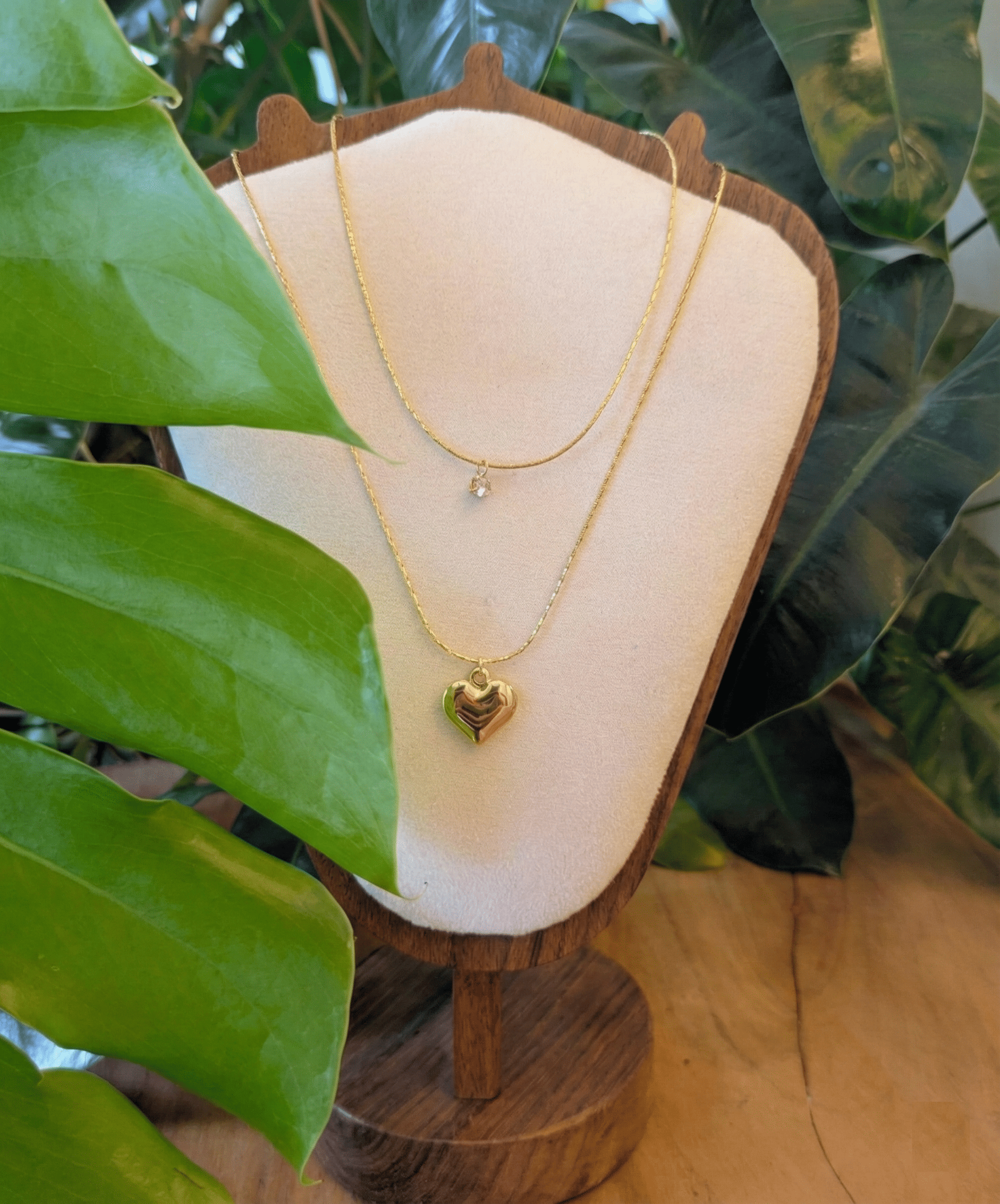 A close-up photograph shows two layered gold necklaces displayed on a light beige bust stand with a dark wood base.