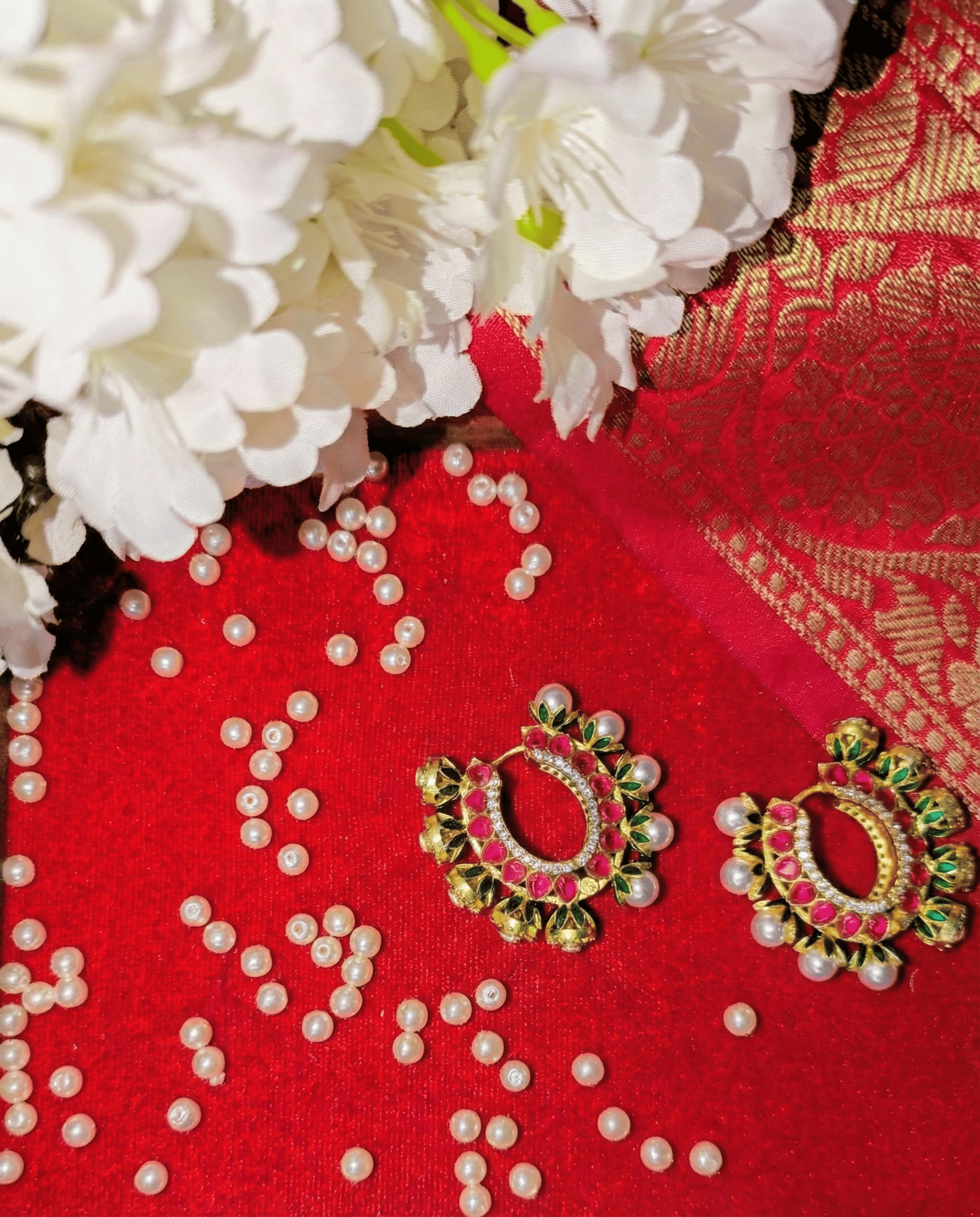 A flat-lay photograph featuring a pair of ornate gold earrings with pink gemstones, green accents, and pearl detailing resting on a vibrant red textured background.