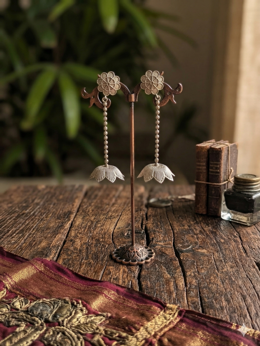 A pair of intricate handcrafted silver dangle earrings on a stylized dark wooden display stand, showcasing beaded chains and floral accents.