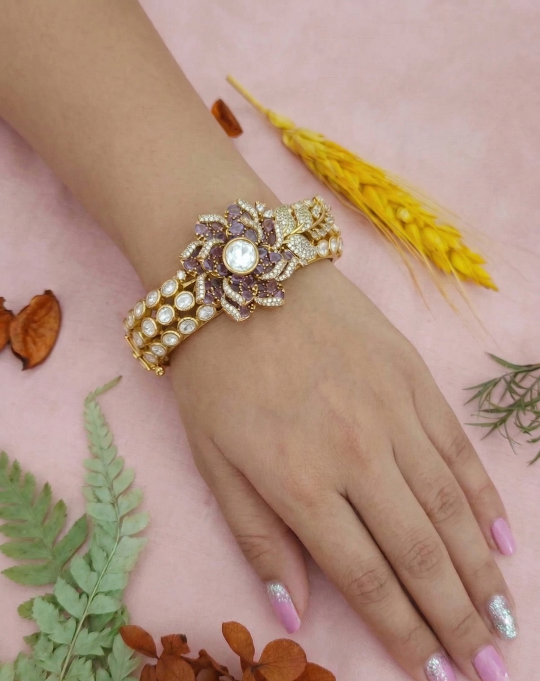 A beautiful hand wearing a traditional Indian Kundan Jadau bangle with sparkling CZ stones and intricate gold-plated filigree work. The bangle is set against a soft pink background with dried botanical elements like fern and wheat.
