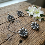 Load image into Gallery viewer, A pair of oxidized silver ear cuffs featuring a floral base connected to a fan-shaped cuff by a thin chain, displayed on a rustic wooden surface next to white flowers.
