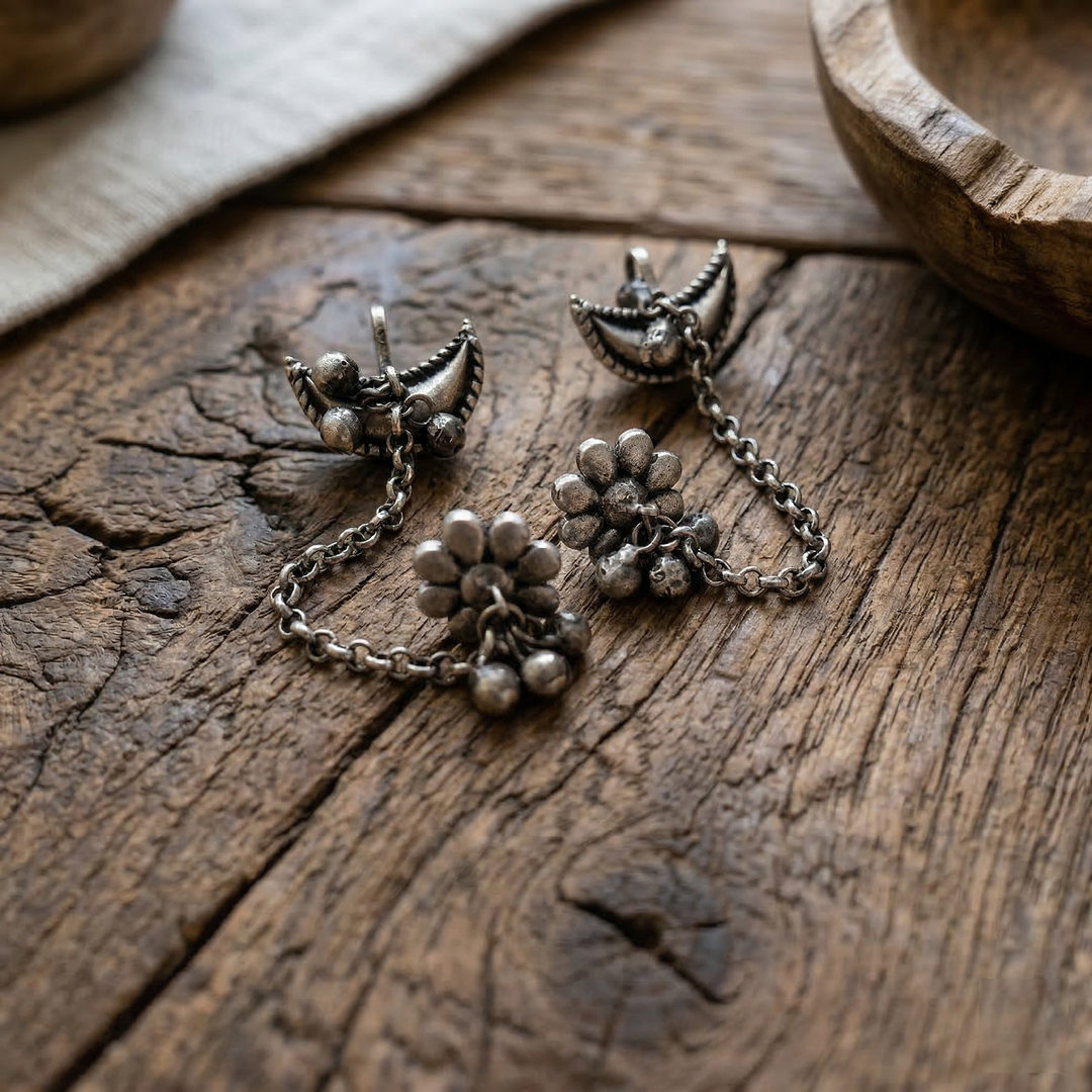 Pair of oxidized silver earcuffs featuring floral motifs, link chains, and small hanging bells on a rustic wooden background.