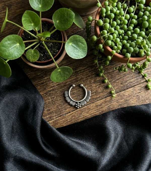 Oxidized silver septum nose ring with detailed tribal design placed on wooden surface with black satin cloth and green potted plants.