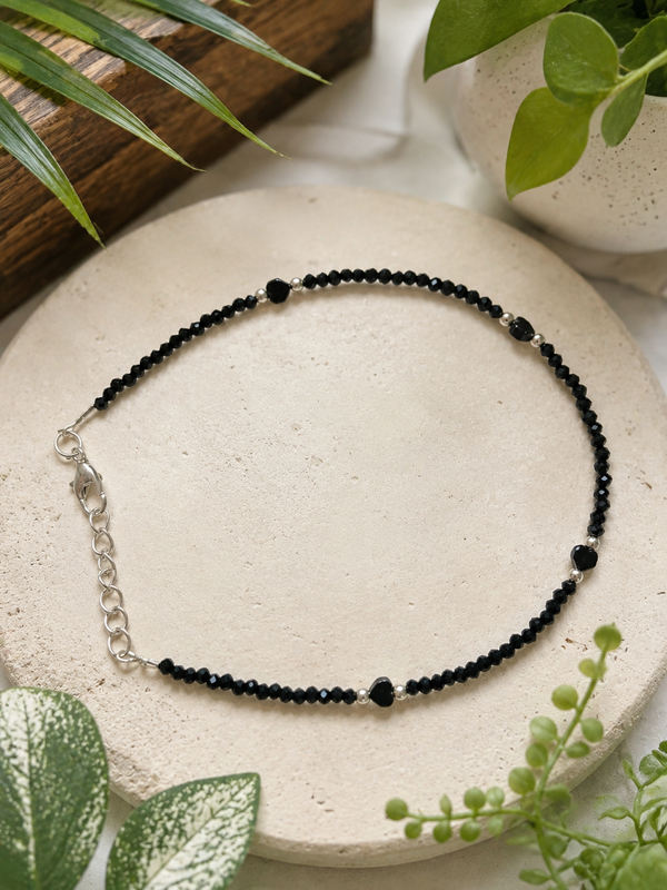 Handcrafted oxidized silver black crystal beaded anklet with adjustable chain, minimalist boho payal for women, elegant everyday silver ankle bracelet displayed on a neutral ceramic surface surrounded by green plants.