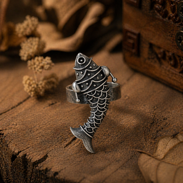 Close-up of an oxidized silver fish statement ring with engraved detailing, placed on a rustic wooden surface with warm earthy tones and dried elements.