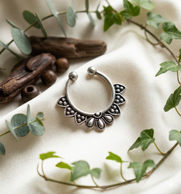 Oxidized silver septum nose ring with floral engraved design placed on soft satin fabric with greenery and wooden accents.